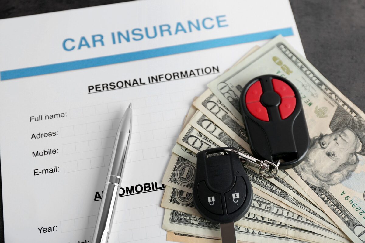 A car insurance application form on a desk with a pen, a set of car keys and a fan of dollar bills.