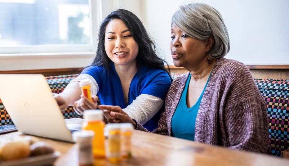 A caregiver points at a laptop screen while holding a pill bottle, next to an older woman with several bottles on the table.