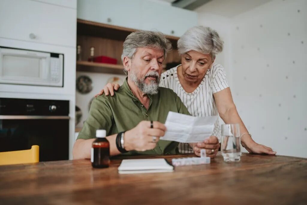 An older couple in a kitchen read a letter together, with a pill organizer and a medicine bottle on the table.