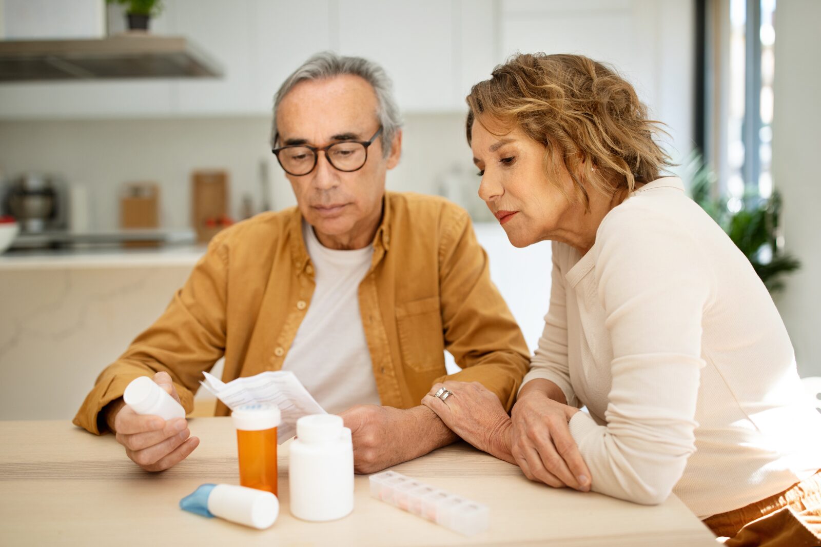 An older couple at a kitchen table read a folded paper beside several pill bottles and a weekly pill organizer.