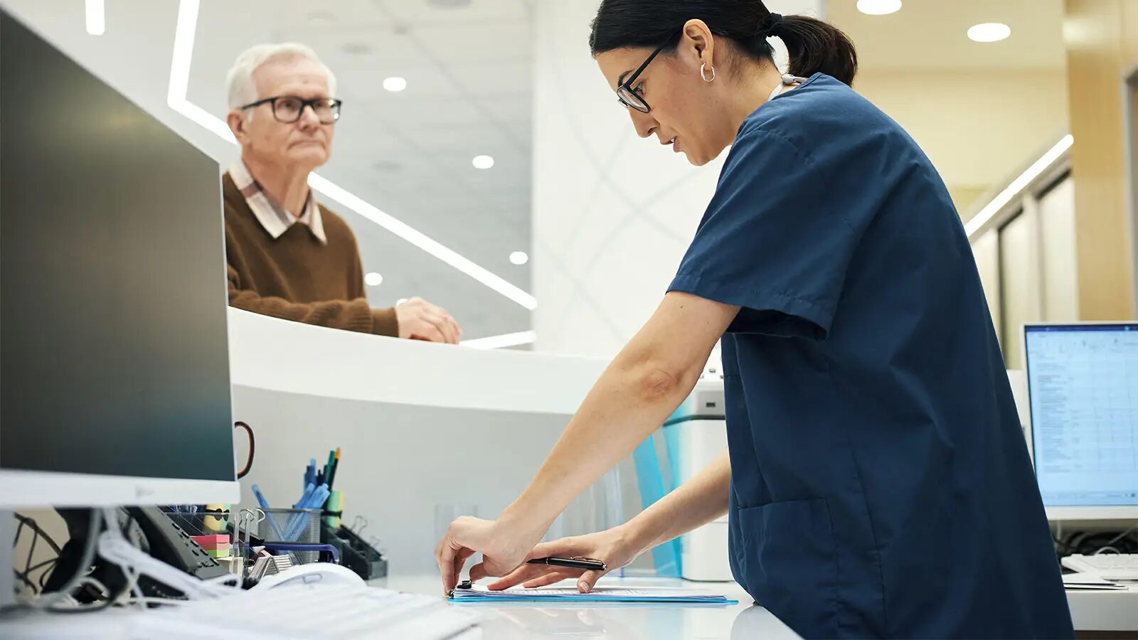 An older man in a brown sweater waits at a clinic front desk while a receptionist in scrubs fills out a form.
