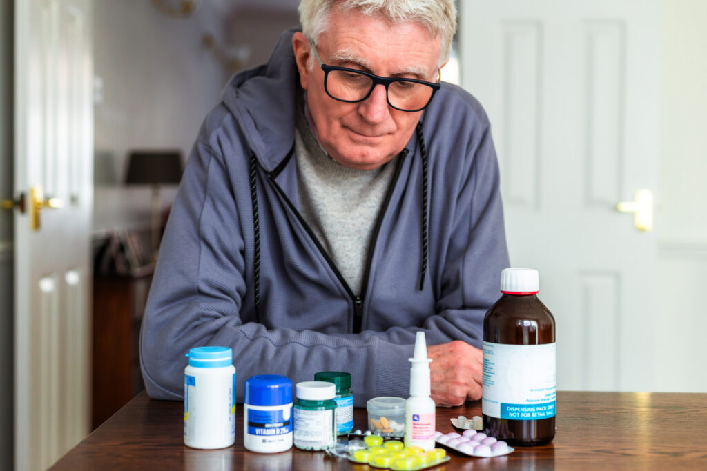 An older man in a grey hoodie looks down at a row of medicine bottles and blister packs on a table.