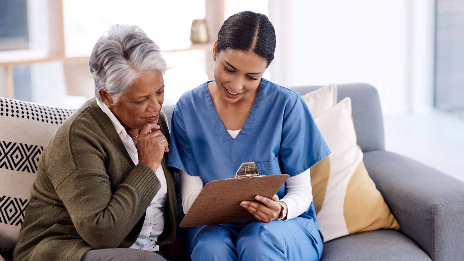 A nurse and an older woman sit on a grey sofa reading a form on a clipboard together.