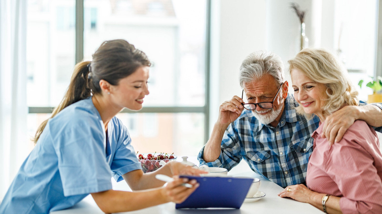 A nurse in blue scrubs goes over a clipboard with an older couple at a kitchen table.