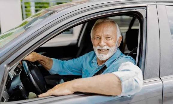 An older man with a grey beard smiling from the driver's seat of his car with the window down.