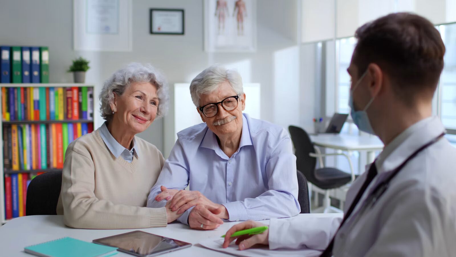 A smiling older couple sit across a desk from a doctor in a white coat who is taking notes.