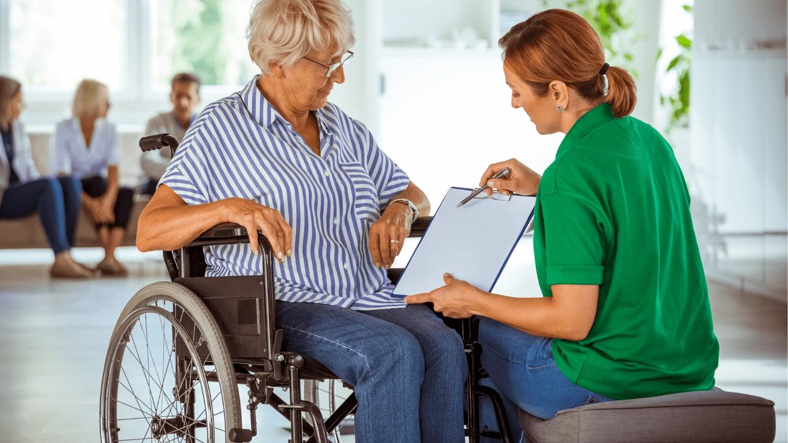 An older woman in a wheelchair reviews a clipboard with a caregiver in a green polo shirt.