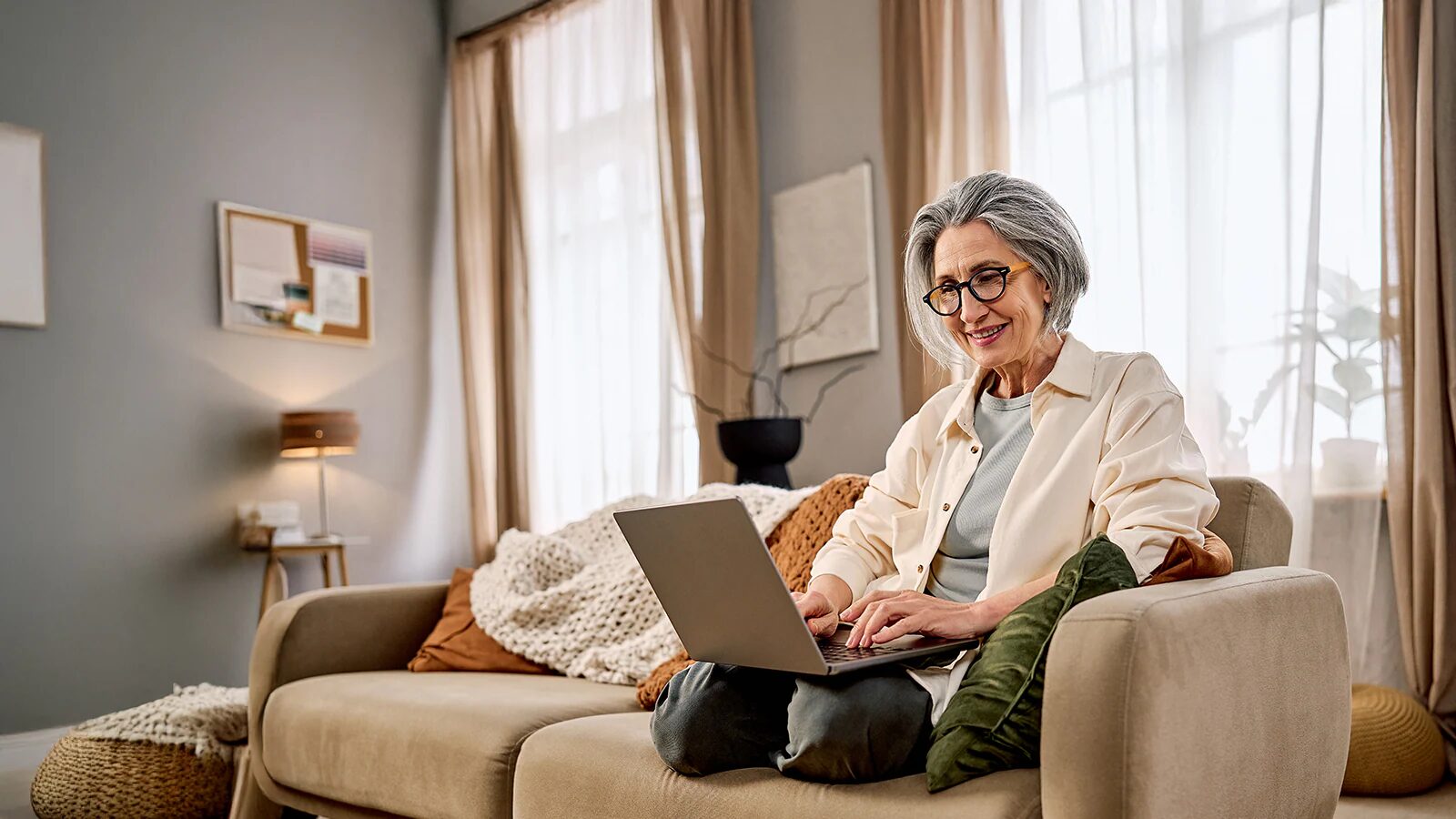 A woman with grey hair and glasses smiles at a laptop on her lap while sitting on a beige sofa.